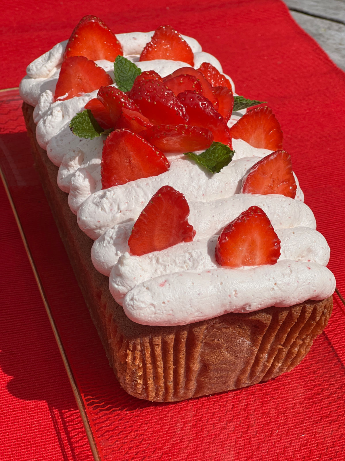 Strawberry Loaf Cake with Strawberry buttercream frosting and fresh strawberries on top. The loaf cake is sat on a glass tray on top of a red cloth in the summer sun.